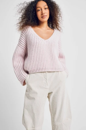 Woman with curly dark hair wearing a light pink ribbed V-neck sweater and pale trousers against a white backdrop.