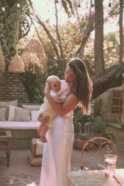 Outdoor portrait of an adult woman holding a baby, set in a sunlit courtyard with lush greenery and rustic furniture, creating a warm family moment.