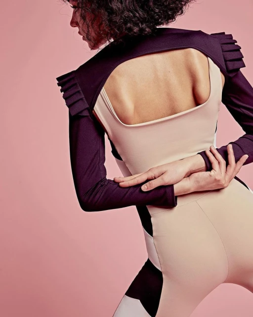 Model in a futuristic beige and black bodysuit with dark purple shoulder pads and sleeves poses in front of a pink background.