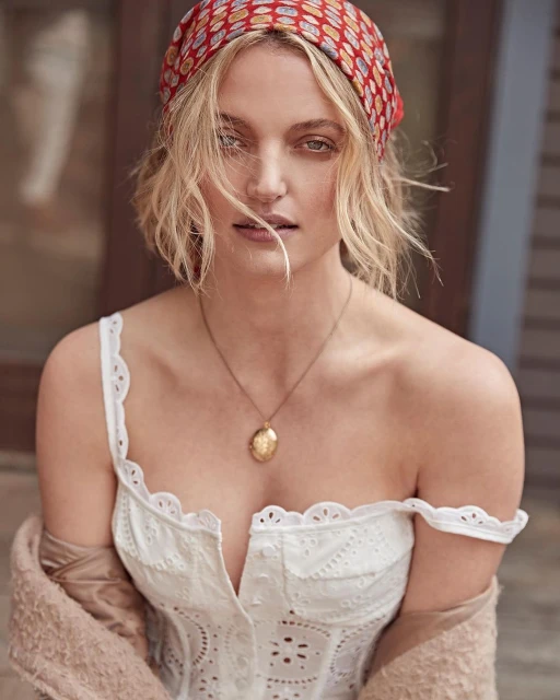 Portrait of a woman with blonde wavy hair wearing a red patterned headscarf and a white top, accentuated by a gold pendant necklace.