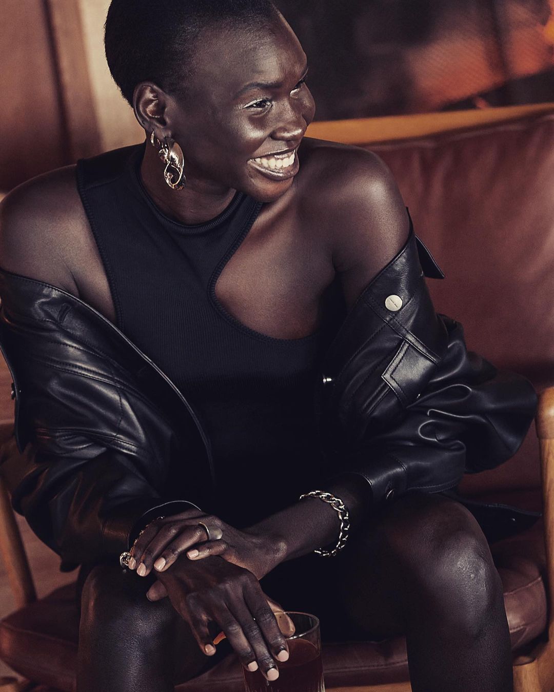 Smiling adult woman in a black dress with leather jacket draped over her shoulder sits indoors with a drink, wearing statement earrings.