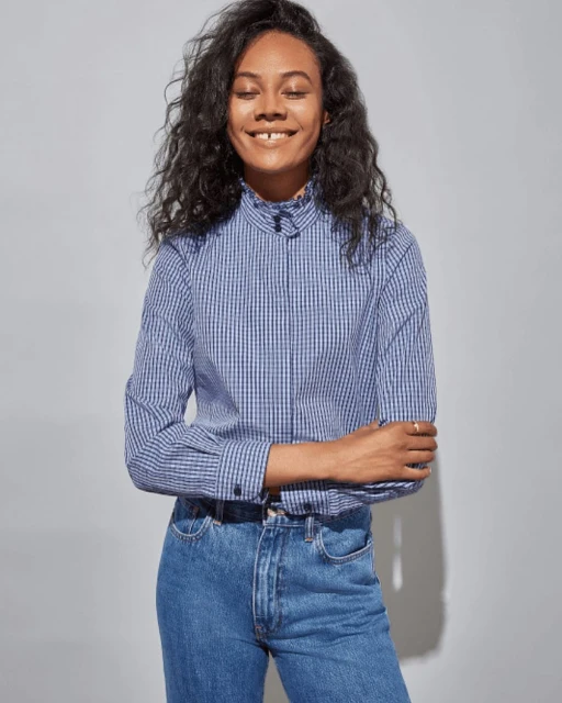 Portrait of a smiling dark-haired woman in a blue plaid shirt and jeans against a gray background in a medium studio shot.