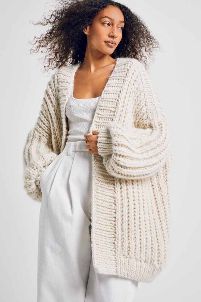 Woman wearing an oversized knit cardigan and white pants in a studio setting. Fashion editorial promoting knitwear.