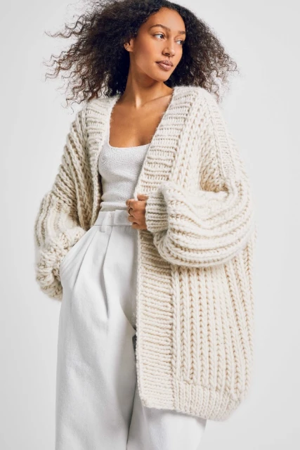 Woman wearing an oversized knit cardigan and white pants in a studio setting. Fashion editorial promoting knitwear.