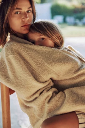 Portrait of a woman holding a child in a cream fleece sweater against a blurred outdoor background