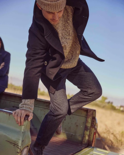 Man in peacoat and beanie climbs into truck bed in an outdoor landscape setting on a bright day. Second person in background.