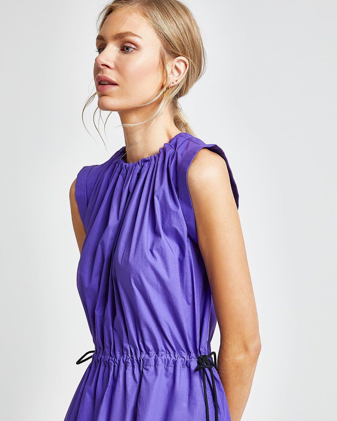 Blonde woman in a violet dress poses in a studio with a neutral background. The dress has a cinched waist and draped neckline.