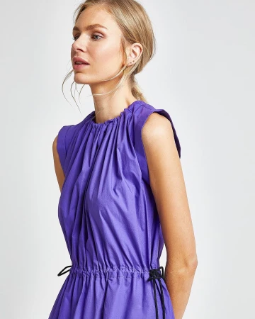 Blonde woman in a violet dress poses in a studio with a neutral background. The dress has a cinched waist and draped neckline.
