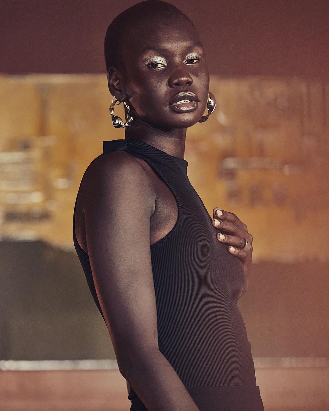 Portrait of an adult woman with a shaved head wearing a black top and silver hoop earrings, set against a blurry neutral background.