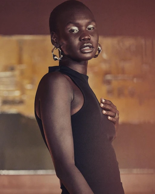 Portrait of an adult woman with a shaved head wearing a black top and silver hoop earrings, set against a blurry neutral background.