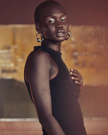 Portrait of an adult woman with a shaved head wearing a black top and silver hoop earrings, set against a blurry neutral background.