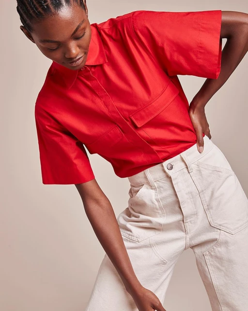 Adult woman poses wearing a red collared shirt with pockets and wide-leg beige pants indoors against a neutral background.