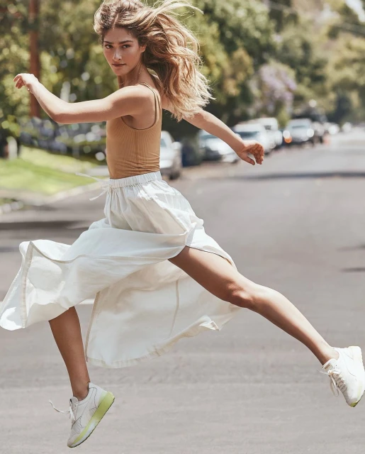 Woman in motion, captured mid-jump on a tree-lined street, wearing a tan top, white skirt, and matching sneakers with neon soles.