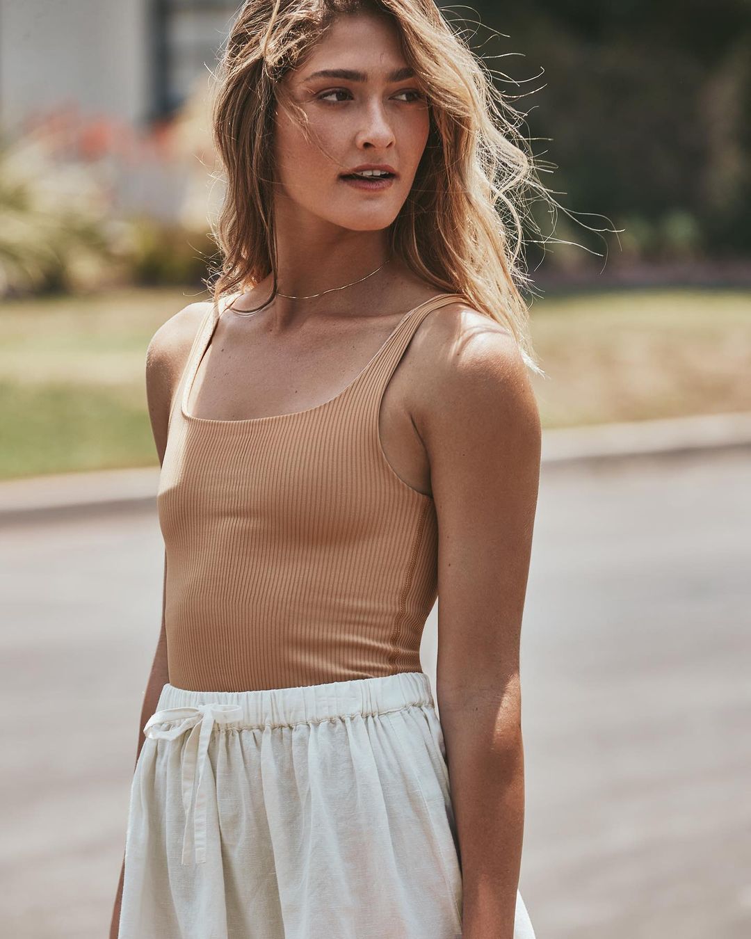 Woman in a ribbed tan bodysuit and white drawstring shorts stands outside with soft light and bokeh from green foliage in background.