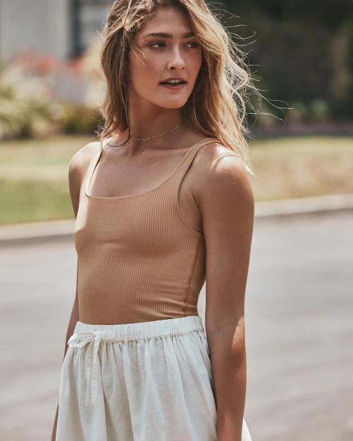 Woman in a ribbed tan bodysuit and white drawstring shorts stands outside with soft light and bokeh from green foliage in background.