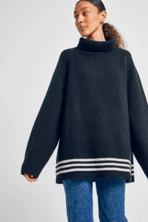 Woman wearing an oversized dark turtleneck sweater with white stripes and blue jeans against a white background