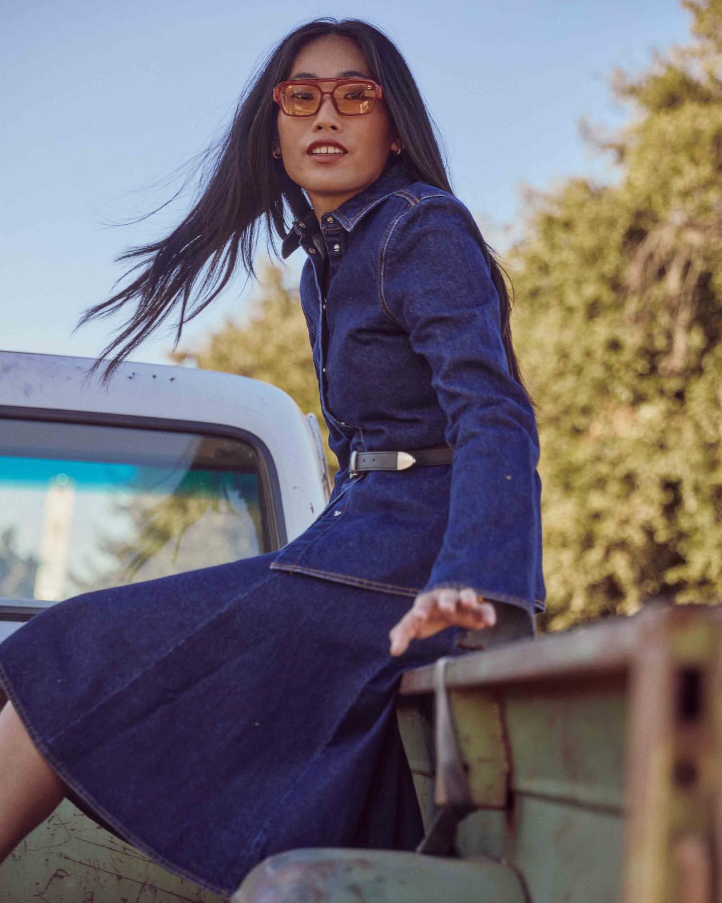 An Asian woman in sunglasses and a denim dress sits on the back of a vintage pickup truck with trees and sky in the background.