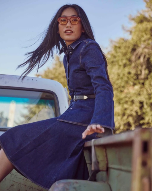 An Asian woman in sunglasses and a denim dress sits on the back of a vintage pickup truck with trees and sky in the background.