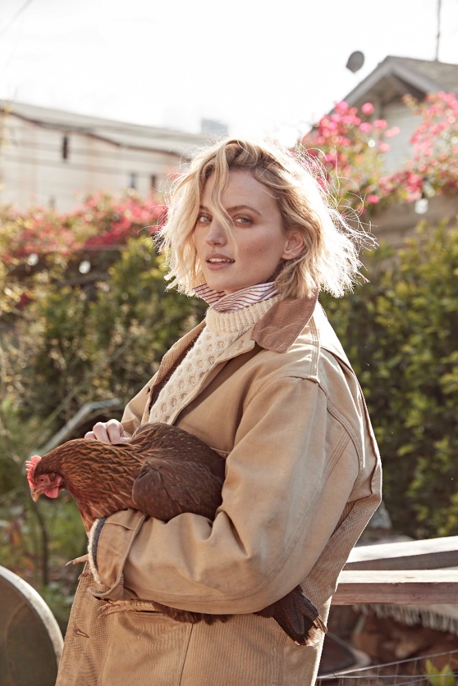 Woman in a tan coat stands outdoors holding a brown chicken with blooming red flowers and a building in the background on a sunny day.