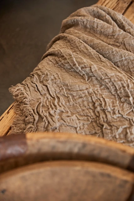 Textured brown fabric drapes across weathered wood surface with worn edges and a rustic aesthetic. Light and shadow accentuate the textile.