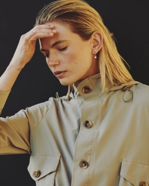 Woman in neutral-toned coat poses indoors. Close-up showcases her face, wet hair, and the coat's buttons against a dark background.