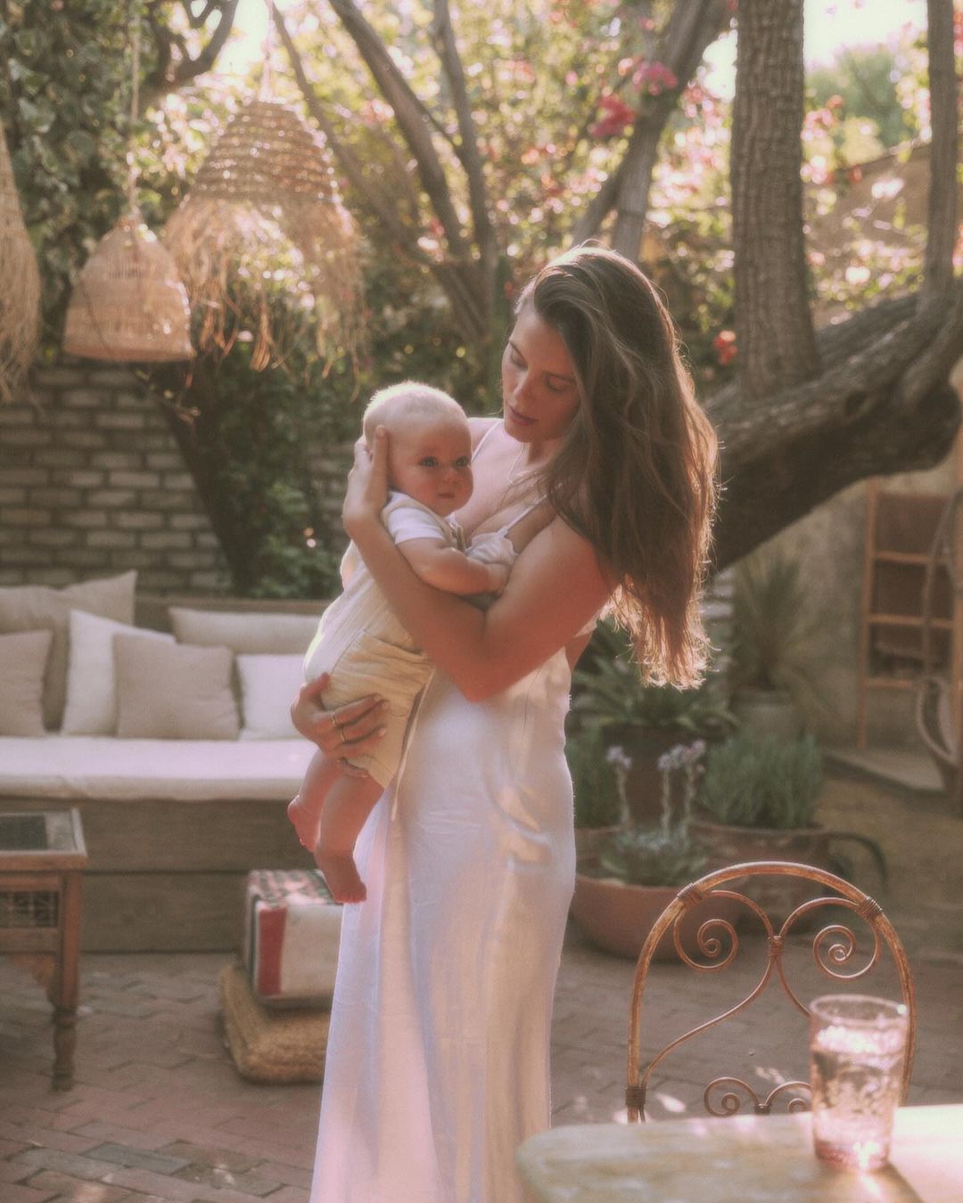 A mother gently holds her baby outdoors in soft light, showcasing a tender family moment with neutral tones and natural textures.