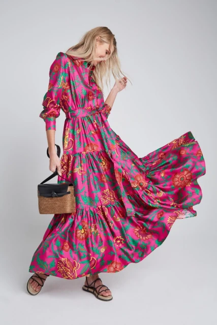 A blonde woman poses in a bright pink floral dress with a black and tan handbag, and strappy sandals against a plain backdrop.