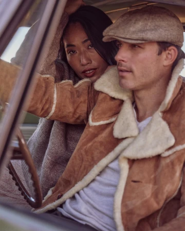 Man wearing a shearling jacket and flat cap sitting in a car alongside a woman leaning from the back seat.
