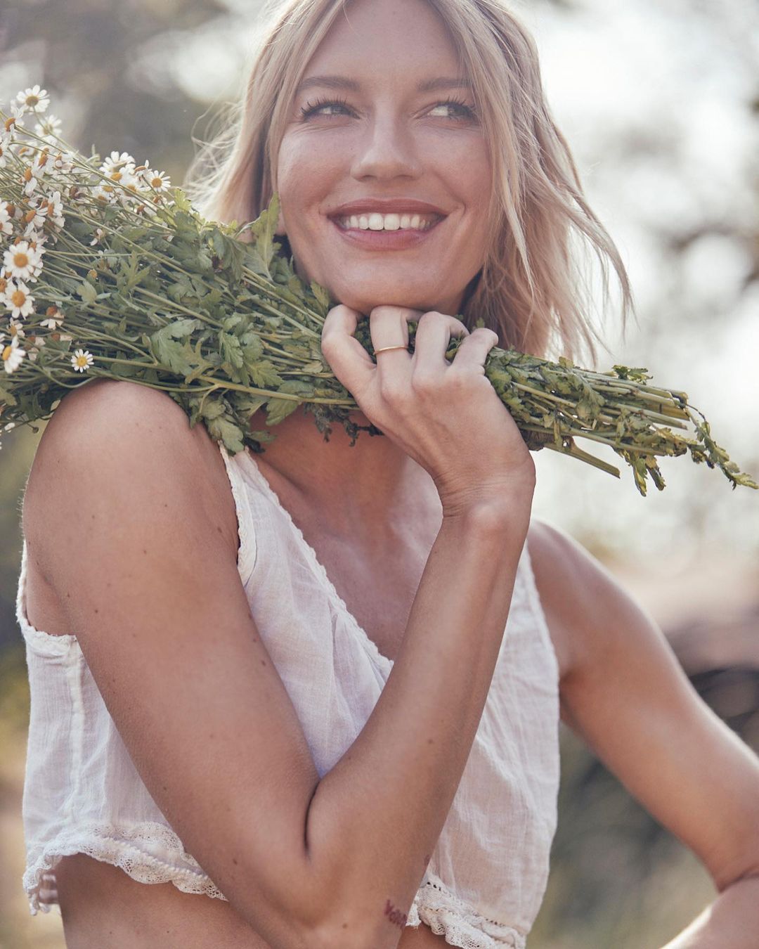 Smiling woman in white top holds chamomile bouquet outdoors. Blonde hair, natural light, and soft focus create a serene atmosphere.