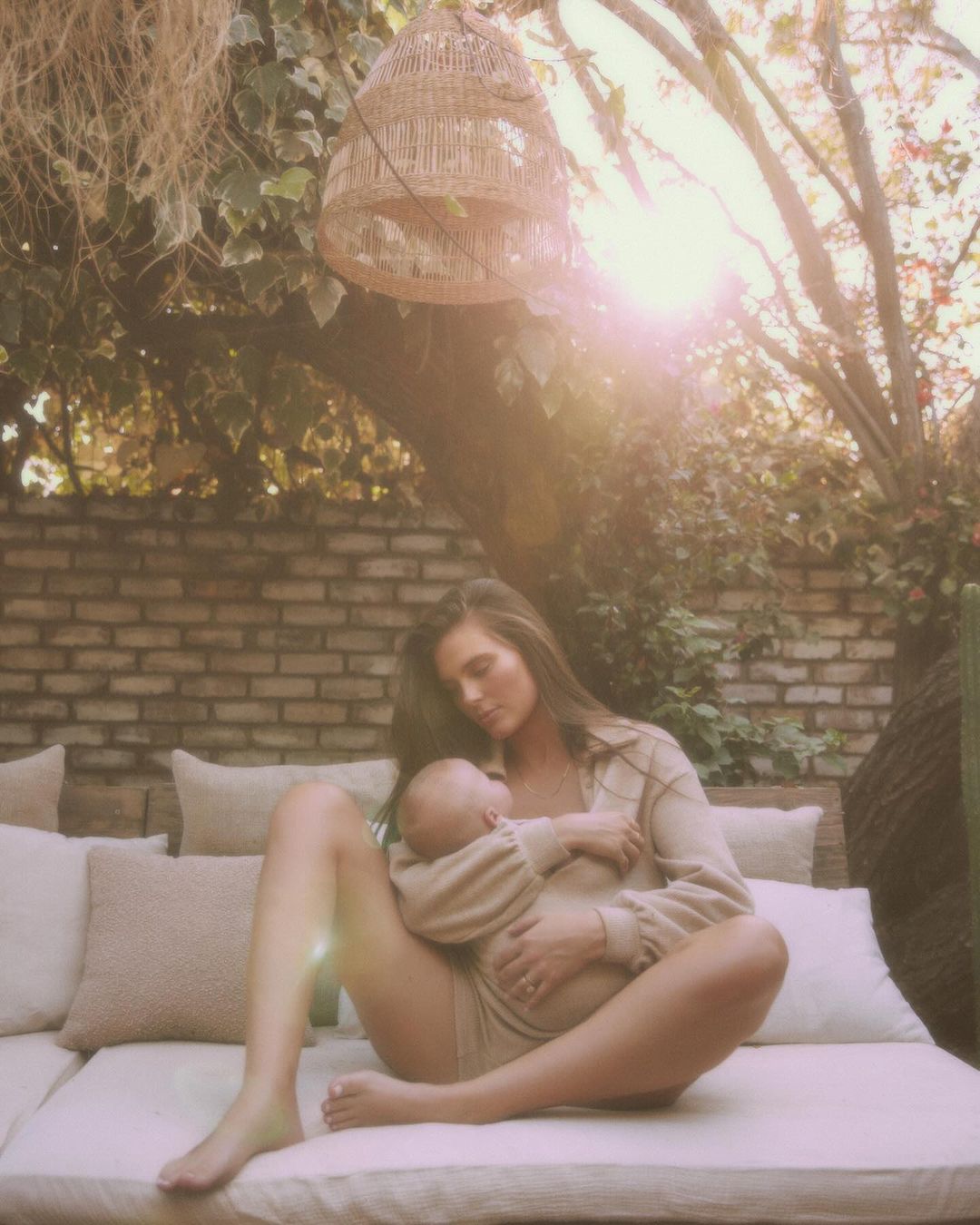 Woman sits on cushions outdoors holding a baby. They both wear matching beige outfits with a brick wall and leafy tree in the background.