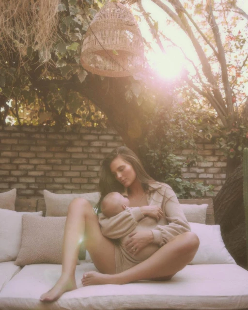 Woman sits on cushions outdoors holding a baby. They both wear matching beige outfits with a brick wall and leafy tree in the background.
