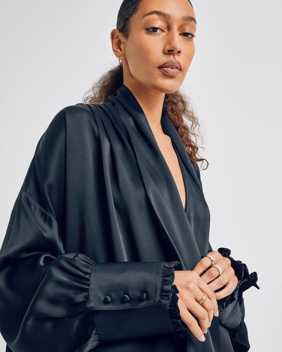 Close-up of an adult female wearing a silky black robe with gold rings, set against a white backdrop, highlighting a blend of fashion and elegance.