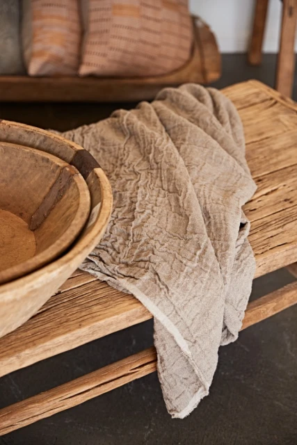 Still life of a rustic wooden bowl and textured beige linen cloth on a wooden bench, with soft lighting and a neutral color palette.