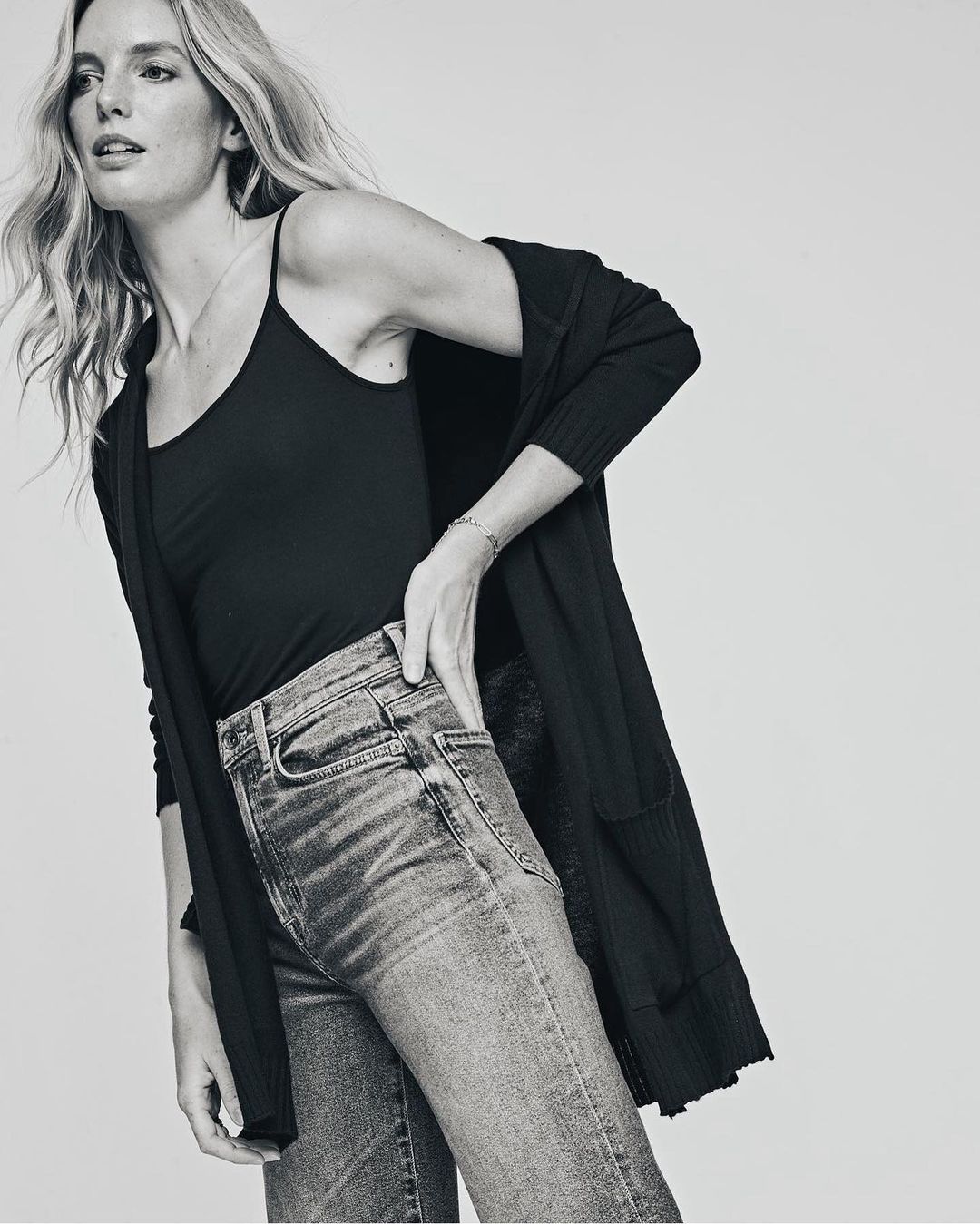 Woman in a black camisole, jeans, and cardigan poses in a studio with a hand on her hip. Monochrome fashion shot.
