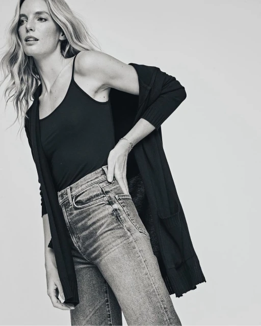 Woman in a black camisole, jeans, and cardigan poses in a studio with a hand on her hip. Monochrome fashion shot.