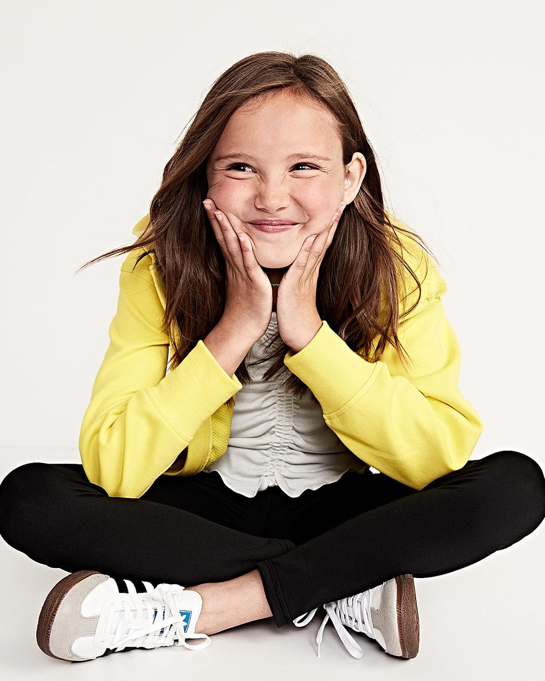 Smiling young girl in a yellow jacket poses in a studio with a white backdrop. She is sitting cross-legged and smiling with her hands on her face.