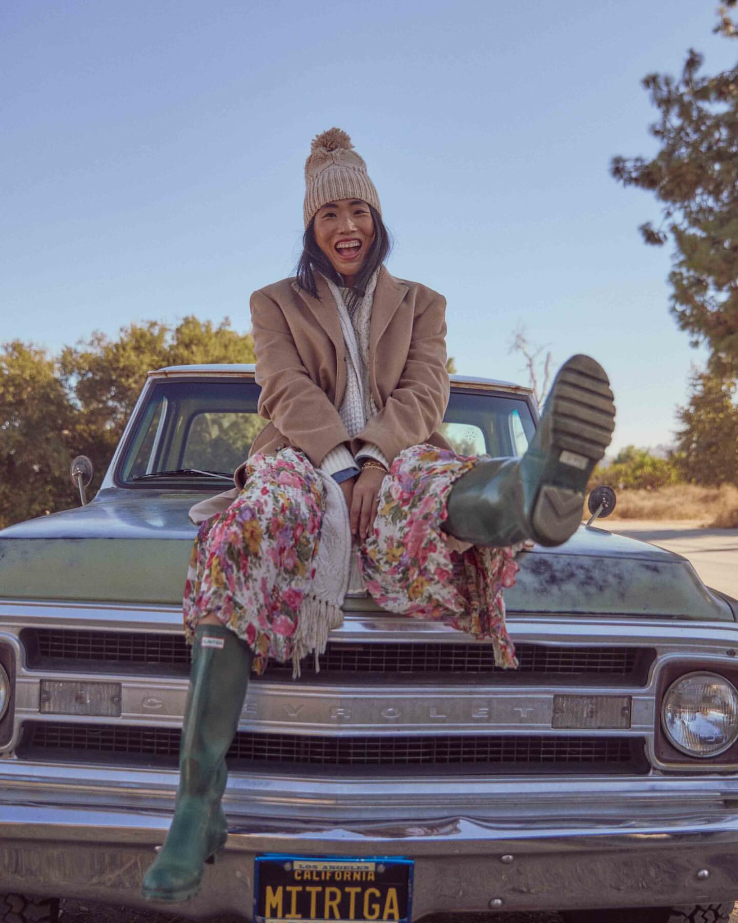 Woman in a floral dress and rubber boots sits on the hood of a vintage truck, smiling, in an outdoor setting with trees and blue sky.