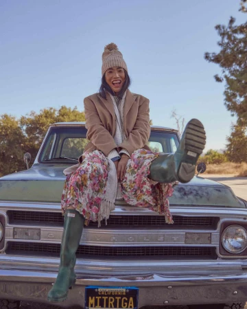 Woman in a floral dress and rubber boots sits on the hood of a vintage truck, smiling, in an outdoor setting with trees and blue sky.