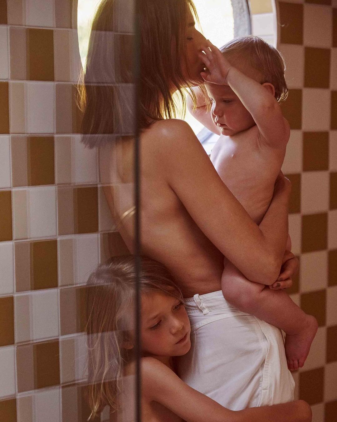 A mother standing indoors, partially obscured by glass, holding a toddler and being hugged by a young child in warm lighting.