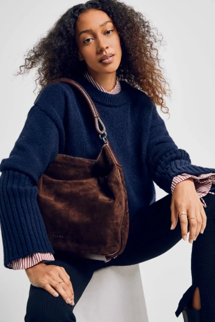 Adult woman poses with brown suede shoulder bag in a studio setting. She wears a navy sweater and black leggings.