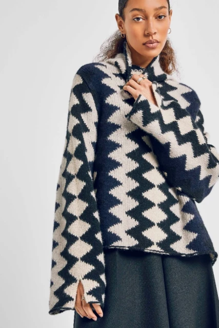 Woman in a navy and beige patterned sweater with wide sleeves, standing indoors. The sweater features a high neck and zigzag patterns.