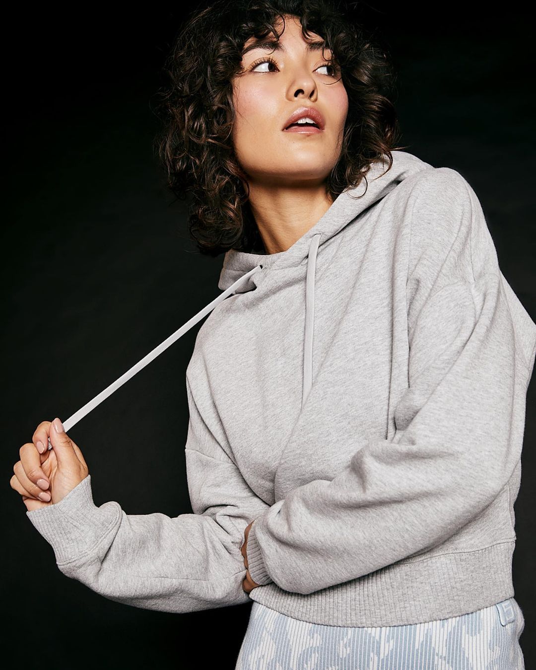 Woman in a grey hooded sweatshirt poses against a dark background. She is looking up and away from the camera with a neutral expression.
