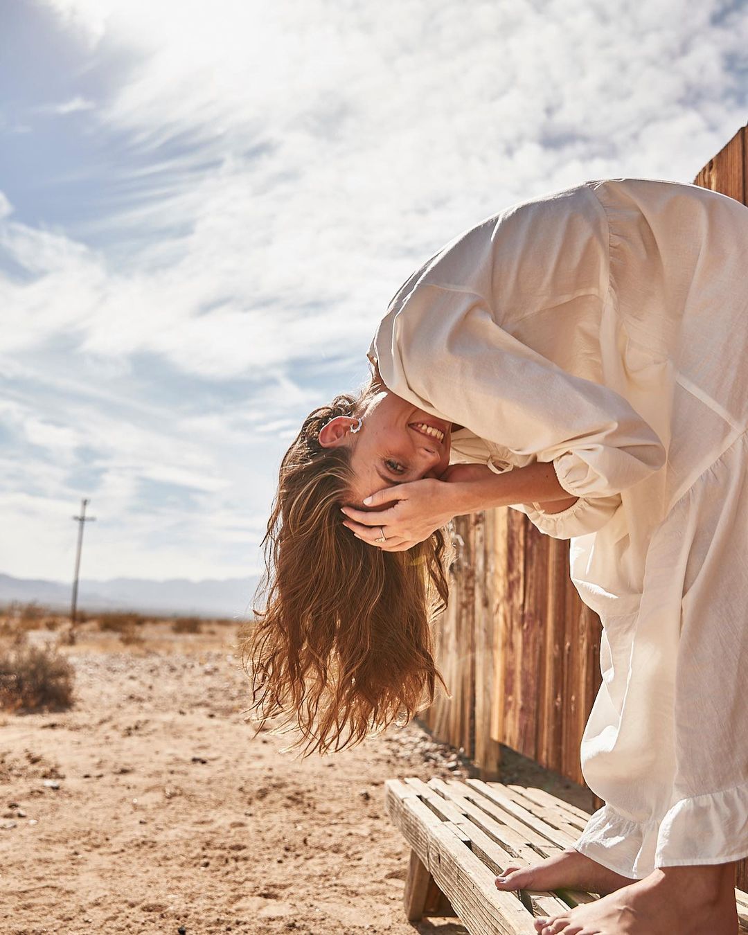 A smiling young woman bends over a rustic bench in a desert setting, wearing a white dress, her long hair falling towards the ground.