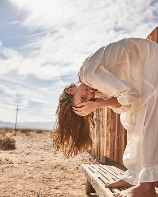 A smiling young woman bends over a rustic bench in a desert setting, wearing a white dress, her long hair falling towards the ground.