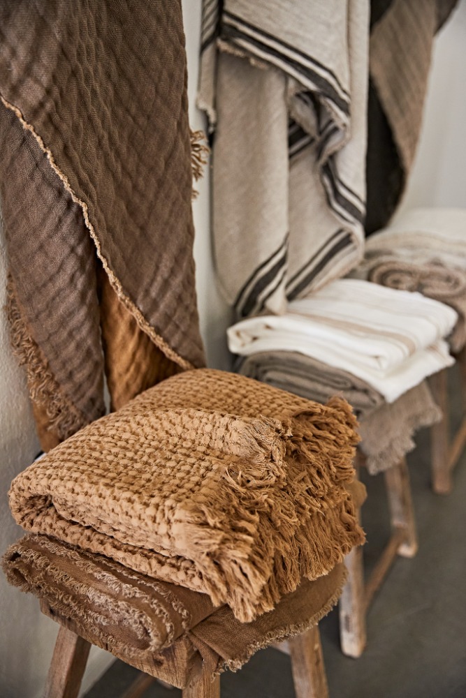 Stacks of folded and draped textile throws and blankets in neutral colors displayed on vintage wooden stools in a retail setting.