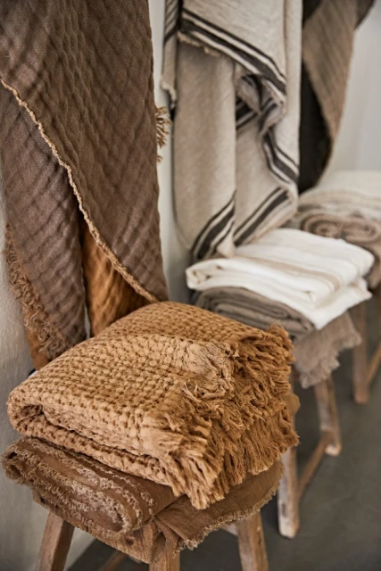Stacks of folded and draped textile throws and blankets in neutral colors displayed on vintage wooden stools in a retail setting.