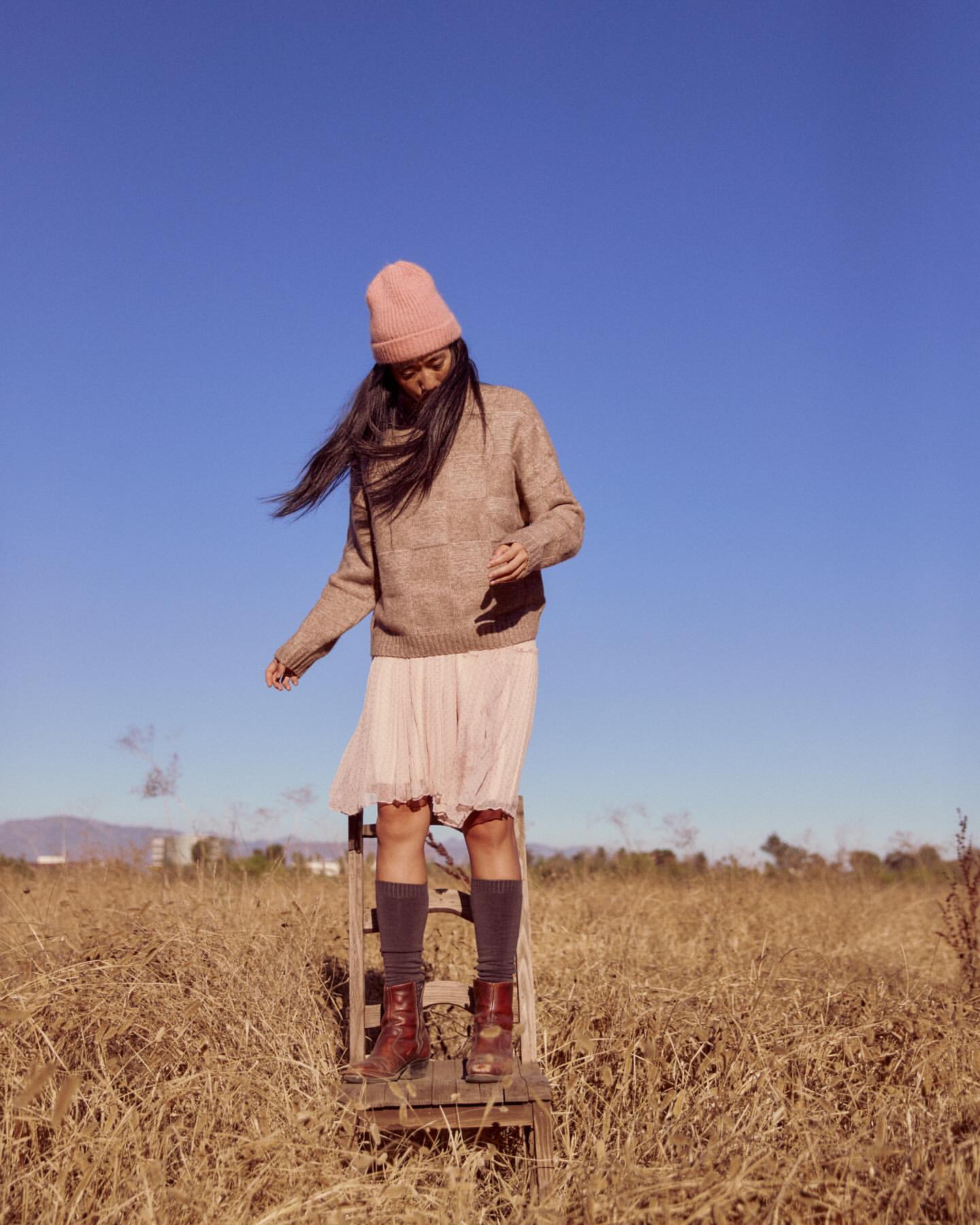 Young adult woman in a pink hat and neutral sweater standing on a wooden chair in a field under a clear blue sky in a fashion shot.
