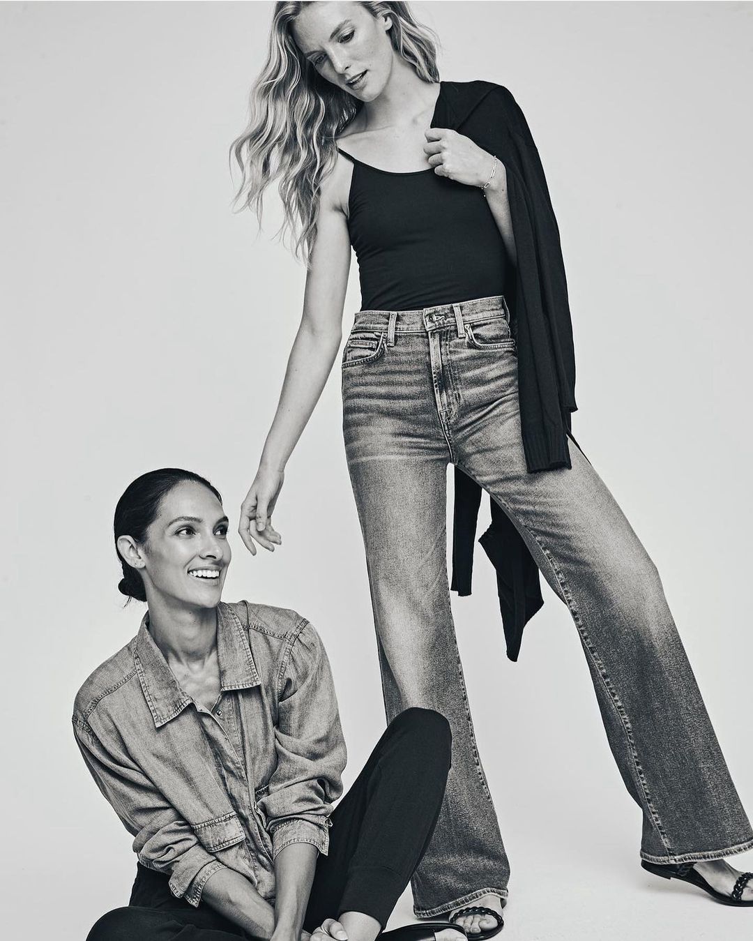 Two women posing indoors in a black and white fashion shot. One woman stands wearing jeans, a black top and a jacket. The other sits smiling.