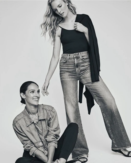 Two women posing indoors in a black and white fashion shot. One woman stands wearing jeans, a black top and a jacket. The other sits smiling.