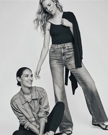 Two women posing indoors in a black and white fashion shot. One woman stands wearing jeans, a black top and a jacket. The other sits smiling.